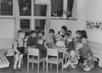 XXII-133-01-2 Kleuterschool aan de Libellenstraat met interieur: kinderen rondom een begeleider tijdens speeltijd.