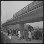 22371-4-11 Groep mensen rond het onthulde beeld 'De Dijkwerker'; daarboven rijdt een metrorijtuig over het viaduct.
