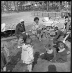 7024 Spelende kinderen in speeltuin en zandbak; begeleidsters en kinderwagens.