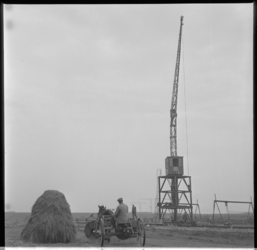 788-3 Boer rijdt op landbouwwerktuig, voortgetrokken door een paard, tussen een hooiberg en een hijskraan.