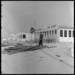 THO-1315 Een man staat op het strand bij houten balken voor de opbouw van strandpaviljoens. Op de achtergrond paviljoen ...
