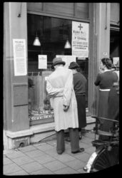 1980-5599 Mensen lezen proclamaties en bekendmakingen over de bevrijding in de etalageruit van een winkel. In de ...