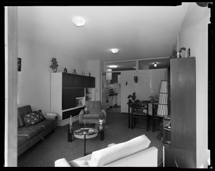 L-10055 Woonkamer van een flatwoning. In de kamer staan stoelen, een eettafel, ee salontafel en een lamp. Links staat ...