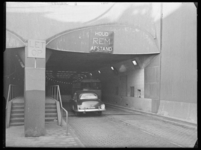 L-1058 Een passagiersbus en een auto rijden de Maastunnel in. Boven de ingang hangen borden met 'Houd Rem Afstand' en ...