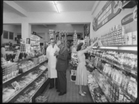 L-3627 Vrouw doet boodschappen met een winkelmandje in een winkel met levensmiddelen aan de Meent. Links van haar ...