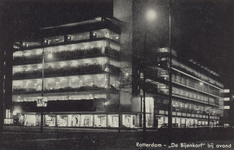 PBK-1266 Het warenhuis De Bijenkorf aan de Schiedamse Vest bij avond met verlichte etalages, gezien uit het noordwesten.
