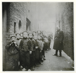 1975-1475 Kinderen wachtend in een rij, gevormd in een poort onder leiding van een politieman.