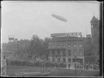 1976-4194 Zeppelin boven Rotterdam. Een menigte mensen op de Schiekade kijkt naar de Zeppelin. Rechts het gebouw van ...