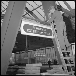 10192 Man op ladder onteert bord met tekst 'Schiedam Rotterdam West' boven een perron van het spoorwegstation in Schiedam.