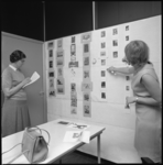 11008 Twee dames verzamelen informatie over prentbriefkaarten die op een wand zijn bevestigd.