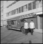 11104-3 Twee brandweerlieden poseren voor een brandweerauto met uitschuifbare ladder.
