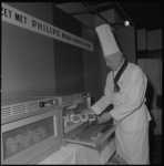 11772 Een kok, met muts op hoofd en onderscheiding om de nek, demonstreert een 'magnetronische oven' van Philips.