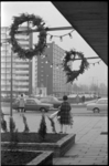 21218-3 Dagfoto van kerstkransen en -lampjes aan winkelluifel; op de achtergrond hoogbouwflats.
