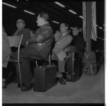 3173-2 Wachtende mannen zitten op stoelen en hebben koffers bij de hand.