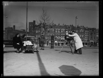 R-1924 Straatfotograaf fotografeert twee vrouwen die op een bank zitten bij hun kinderwagen met baby. De camera op ...