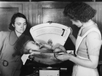1977-3679 Een verpleegkundige van de GGD (Gemeentelijke Geneeskundige Dienst) weegt een baby.