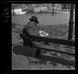 5678 Op het Willemsplein leest een man met hoed een krant. Op de achtergrond liggen schepen aan de kade van de Nieuwe Maas.
