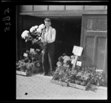 5825 Op de rechter Rottekade staat een jonge verkoper met bloemen in zijn hand voor een opslagplaats met planten.