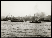 FD-3974 Stoomsleper sleept vrachtschepen met lading op de Nieuwe Maas. Op de achtergrond liggen stoomschepen aan de ...