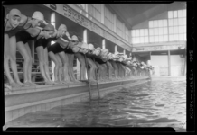 THO-1150 Tijdens een zwemles staat een rij leerlingen in duikhouding aan de rand van het zwembad. Uit een serie over ...