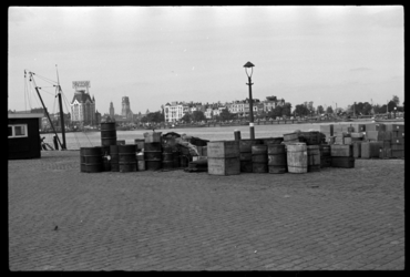 1980-5325 Op de Maaskade staan goederen en tonnen. Op de achtergrond is de Nieuwe Maas en de Rechter Maasoever te zien. ...