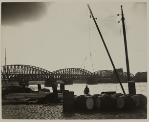 1990-1060 Goederen in tonnen langs de Maaskade. Op de achtergrond de spoorbrug over de Nieuwe Maas.