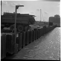 17419 Een met houten palen beladen vrachtwagen staat naast het kanaal dat op het Weena is aangelegd voor de aanleg van ...