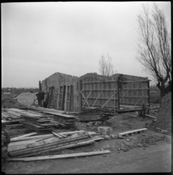 1969 De bouw van noodwoningen in de Wielewaal. Bouwvakkers rusten uit tegen een houten wand, omringd door meer bouwmateriaal.