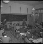 4788 Zicht op een schoolklas in een noodschoolgebouw aan de Van Aersenlaan. Vooraan bij het schrijfbord zit de leraar, ...