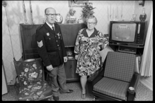 2021-53-7 Een echtpaar staat tussen meubels in hun woning. De man draagt een jas van de brandweer met medailles. Rechts ...