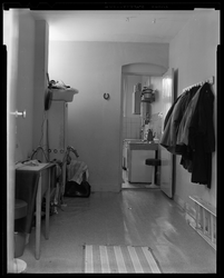 L-10066 De bijkeuken in een woning met daarachter de keuken. In de kamer hangen jassen aan de kapstok, een koelkast en ...