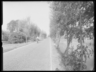 L-1564 Een fietser fietst over de 's-Gravenweg, een landweg langs boerderijen. Aan weerszijden sloten en weilanden.