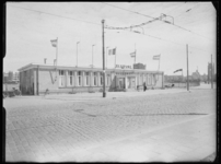 L-1943 De ingang van luchthaven Heliport aan de Hofdijk. Aan de gevel hangen vlaggen. Op de voorgrond het spoor van de ...