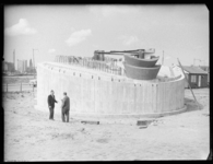 L-2159 Twee mannen overleggen bij het betonnen voetstuk voor het Nationaal Koopvaardijmonument De Boeg. Op de ...