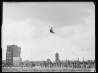 L-257 Een helikopter landt op luchthaven Heliport in Katshoek. Omstanders kijken toe. Links een gebouw met het ...