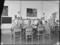 L-2811 Leerlingen zitten in een groep aan tafels in een klaslokaal een kleuterschool aan de Ameidestraat.