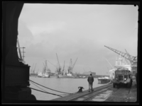 L-4913 Zicht op de Merwehaven. Langs de kade loopt een havenarbeider en geheel rechts staat een vrachtwagen voor de ...