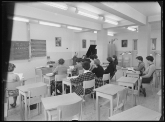 L-537 Lokaal voor muziekles. Een docent zit achter een vleugel, leerlingen luisteren. Op het bord staat notenschrift. ...