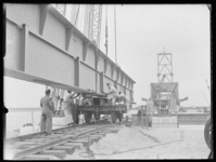 L-766 Bouwvakkers verplaatsen een stalen balk op een treinstel bij de bouw van de Botlekbrug. Op de achtergrond is te ...