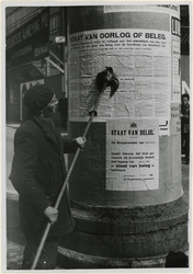 XXXIII-565-01 Een man plakt op een reclamezuil een biljet waarop de staat van beleg wordt aangekondigd.