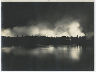 2018-208-1 De stad in in brand tijdens het bombardement van 14 mei 1940, gezien vanaf het water. Op de voorgrond zijn ...