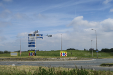 308 Verkeersrotonde met richtingborden bij de G.K. van Hogendorpweg. Op de achtergrond een landend vliegtuig voor ...